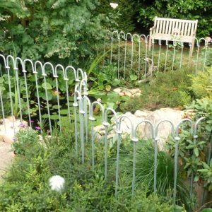 Zaun aus einzelnen verzinkt gebogenen Stäben vor einem Teich im Garten